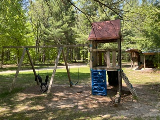A wooden playground set with swings, a blue climbing wall, and an elevated playhouse stands in a grassy, shaded yard surrounded by tall trees under a sunny sky.