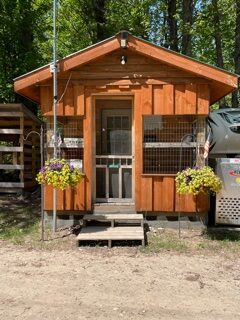 A small wooden cabin with a porch, two hanging baskets of yellow and purple flowers, and a wire enclosure on each side of the front door, surrounded by trees and greenery.