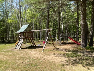 A wooden playground set with swings, slides, and climbing features stands in a grassy, shaded yard surrounded by tall trees on a sunny day.