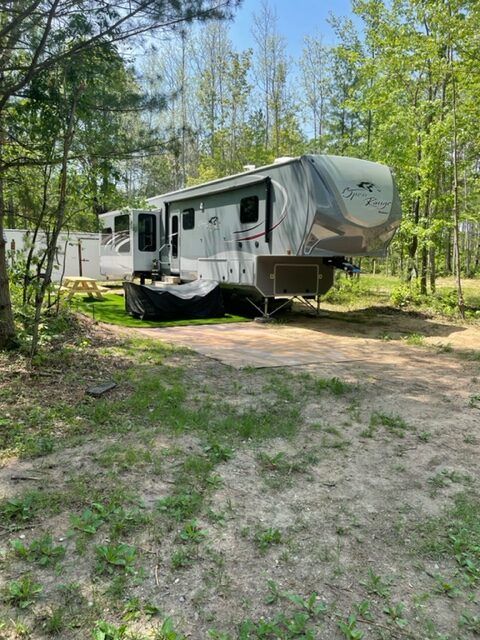 A large fifth-wheel RV is parked on a wooded campsite with trees and greenery surrounding it. The RV has slide-outs extended and is set up on a dirt and grass area with sunlight filtering through the trees.